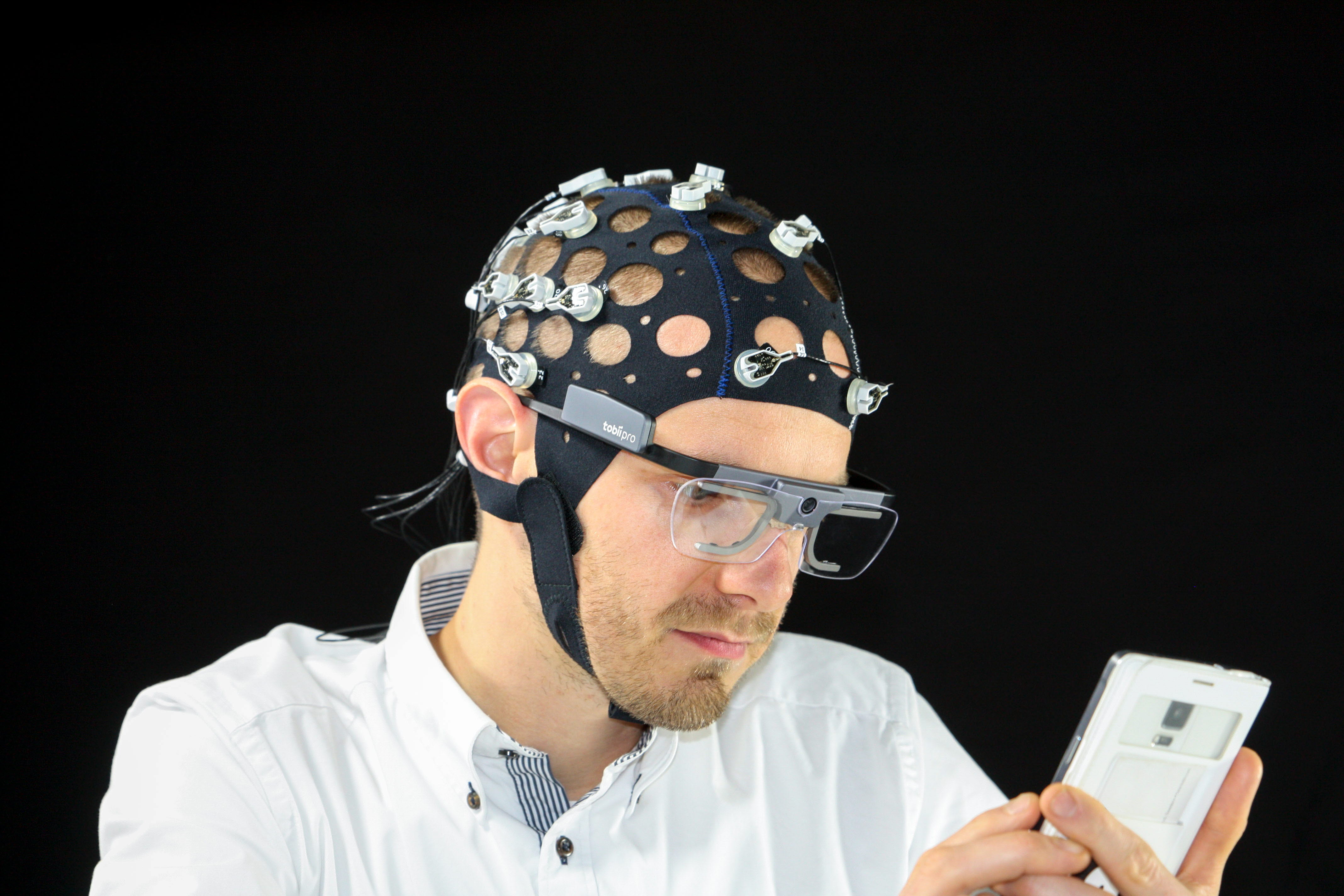A man wearing Tobii Pro Glasses 2 and EEG cap interacts with his mobile phone